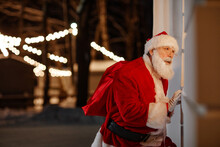 Portrait Of Santa Claus Delivering Gifts Standing Outdoors Peeping Through Window Checking If Family Sleeping