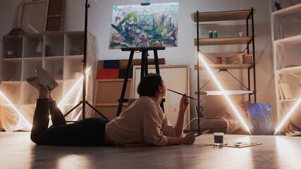 Wall Mural - Creative inspiration. Female painter. Art studio. Thoughtful woman laying floor with palette paints looking around light shadow room interior.