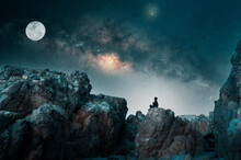Person On The Rock Outdoors Meditating Or Praying At Night Under The Milky Way And Moon
