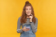indoor studio shot of young ginger female with long red curly hair biting her lip, holding phone with ashamed guilty facial expression, isolated over yellow background