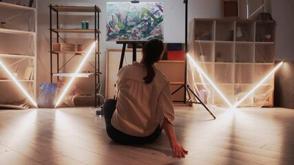 Wall Mural - Artwork creation. Finish process. Art studio. Unrecognizable woman sitting floor enjoying result of abstract painting in dark light room interior.