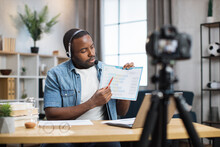 African American Man In Headset Using Laptop And Digital Camera For Online Working Meeting. Male Freelancer Holding In Hand Documents With Charts And Graphs.