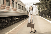 An Elegant Lady In A Dress With An Umbrella And A Vintage Suitcase Is Waiting For Her Train On The Station Platform. A Young Woman In Retro Style Travels By Train.