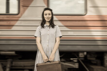 A Young Woman In A Dress And Carrying A Suitcase Is Standing On A Platform With A Blurry Fast-moving Train In The Background. Retro-style Processing. The Concept Of Traveling By Train.