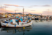 Colorful Fishing Boats Line The Harbor Of The Greek Island Of Aegina, Greece At Dusk, With The Waterfront Promenade And Shops In View.
