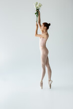 Side View Of Ballerina In Bodysuit Holding Flowers While Standing On Ballet Shoes On Grey