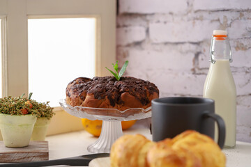 Canvas Print - Chocolate cake next to a window accompanied by milk, orange, a cup of coffee, and croissants