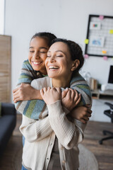 happy african american girl with closed eyes hugging laughing mom at home
