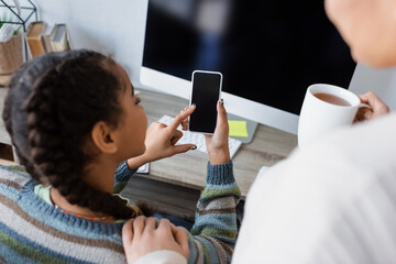 Wall Mural - african american girl using cellphone with blank screen near computer monitor and blurred mom with cup of tea