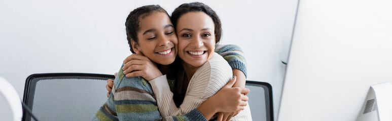 Wall Mural - happy african american woman and teenage girl embracing near computer monitor, banner