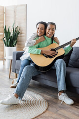 Wall Mural - pleased african american woman looking at camera while hugging daughter playing guitar on sofa