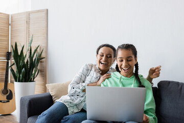 Wall Mural - cheerful african american mother and daughter watching movie on laptop at home