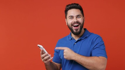 Wall Mural - Young bearded brunet man 20s years old wear blue t-shirt hold use mobile cell phone browsing swipe do online shopping show thumb up loke gesture isolated on plain red orange background studio portrait