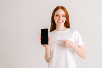 Wall Mural - Studio portrait of positive redhead young woman with wide smile holding blank screen mobile phone and pointing index finger, looking at camera, standing on white isolated background.