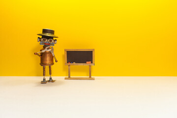Robot teacher professor stands near an empty black chalkboard. Toy mechanical bot and artificial intelligence machine learning concept. copy space on yellow white background