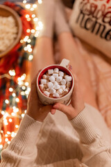 Wall Mural - cup of cocoa or coffee drink with marshmallows in female holding hands over the legs of woman in knitted sweater relaxing and enjoyment the holidays in cozy bed at home on christmas or new year eve