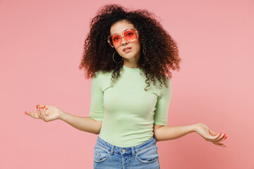 Poster - Puzzled young curly latin woman 20s years old wears mint t-shirt sunglasses spread hands shrugging shoulders have no idea nothing to say isolated on plain pastel light pink background studio portrait.