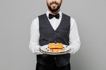 Wall Mural - Young happy barista male waiter butler man wear white shirt vest elegant uniform work at cafe hold cheesecake waffles isolated on plain grey background studio portrait. Restaurant employee concept