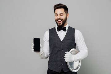 Canvas Print - Happy young barista male waiter butler man 20s wear white shirt vest elegant uniform work at cafe hold mobile phone with blank screen isolated on plain grey background. Restaurant employee concept