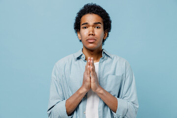 Pleading young black curly man 20s years old wears white shirt hands folded in prayer gesture begging about something making wish asking isolated on plain pastel light blue background studio portrait.