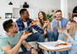 Relaxed multiracial friends having home party, eating pizza in living room, chatting, laughing, spending time together