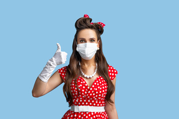 Wall Mural - Protection against coronavirus. Pinup woman in retro dress wearing face mask to prevent covid-19, showing thumbs up