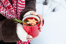 Mulled Wine In Red Cup In Female Hands. Hot Winter Drink With Wine, Spices And Fruit Outdoor On Snowy Background. Copy Space.