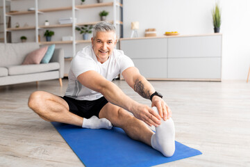 Wall Mural - Sport at home during quarantine. Senior man doing stretch exercises for legs sitting on mat in living room