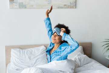 sleepy african american woman in pajamas adjusting sleeping mask and stretching while smiling in bedroom