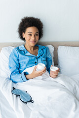 Wall Mural - cheerful african american woman in pajamas holding container with cosmetic cream in bed