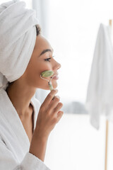 Wall Mural - side view of pleased african american woman in bathrobe and towel massaging face with jade roller in bathroom