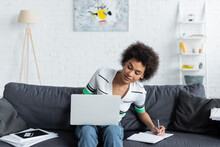 Curly African American Woman Looking At Laptop And Writing In Notebook In Living Room