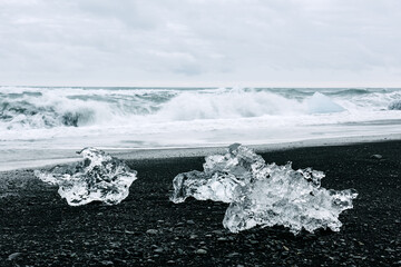 Wall Mural - Iceberg pieces on black Diamond beach near Jokulsarlon lagoon, Iceland. Atlantic ocean waves and cloudy sky on background. Landscape photography