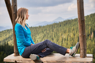 Female traveler spending time on wooden hanging bench and looking at beautiful mountain scenery, thinking about future mountain walking. Pleasant rest in fresh air.