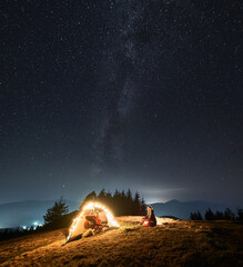 Wall Mural - Male tourist holding guitar in hands recalsing song for his wife, being in tent in their mountain campsite. Romantic atmosphere of sky lit by stars, Milky Way and light from city under hills far away.