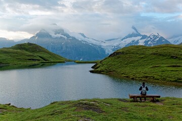 Wall Mural - Tourist sitting on a bench by the grassy lakeside of Bachalpsee & enjoying the panoramic view of majestic alpine mountains under moody cloudy sky in First, Grindelwald, Bernese Highlands, Switzerland