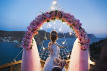 The Bride Holds The Groom's Hand Against The Background Of A Wedding Arch With Bright Lights At Sunset On The Seashore. The Wedding Ceremony.