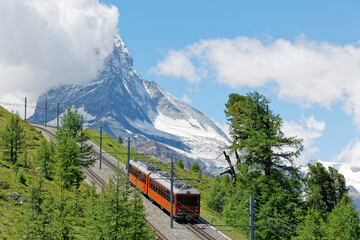 Summer scenery of a cogwheel train of Gornergrat railway traveling thru a forest on the hillside with majestic Matterhorn mountain peak veiled by clouds under blue sky, in Zermatt, Valais, Switzerland