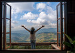 back of Asian woman with freedom arms open standing behind large wooden window with mountain view and cloudy blue sky at Doi Chang, travel attraction in Chiang Rai, Thailand   