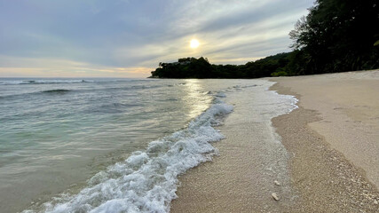 Beautiful sunset at the sea coast. Waves, white foam. Sunshine, reflection of the sun ray on the surface of the water. Sand deserted beach. Sandy shore. Promontory covered by green forest and houses. 