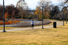 A Woman Jogging On A Smooth Footpath With A Stunning Autumn Landscape In The Park Surrounded By Autumn Colored Trees And Grass With Tall Black Lamp Posts At Centennial Park In Nashville Tennessee USA