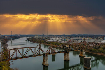 an aerial shot of a rusty iron rail road bridge over the silky green waters of the Cumberland River surrounded by buildings in the cityscape with powerful clouds at sunset in Nashville Tennessee USA 
