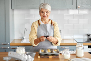 Wall Mural - cooking, food and culinary concept - happy smiling senior woman with smartphone taking picture of cupcakes in baking mold on kitchen at home