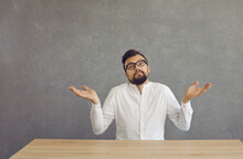 Confused Young Bearded Man Shrugs And Spreads His Arms With A Dumb And Puzzled Expression. Man Is Sitting At A Table On A Gray Background And Does Not Want Or Can Not Perform The Task Assigned To Him