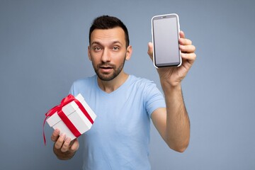 Photo of handsome young unshaven brunet man with beard wearing everyday blue t-shirt isolated over blue background holding mobile phone with blank black screen display for mockup and white gift box