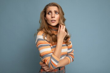 Wall Mural - Portrait photo of young pretty beautiful attractive scared asking thinking blonde woman with wavy-hair wearing striped sweater isolated over blue background with empty space for text