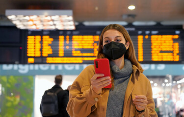 Wall Mural - Traveler woman wearing KN95 FFP2 face mask at the subaway station. Young woman with behind timetables of departures arrivals checking information on her mobile phone inside the train station.