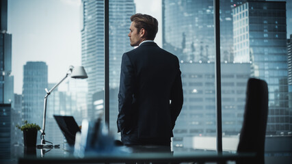 Thoughtful Young Businessman in a Perfect Tailored Suit Standing in His Modern Office Looking out of the Window on Big City with Skyscrapers. Successful Finance Manager Planning Project Strategy.