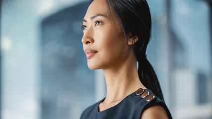 Beautiful Portrait of an Asian Businesswoman in Stylish Black Dress Posing Next to Window in Big City Office with Skyscrapers. Confident Female CEO Smiling. Successful Diverse Business Manager.