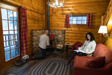 Couple Relaxing With Book And Starting Fire In Cozy Winter Log Cabin